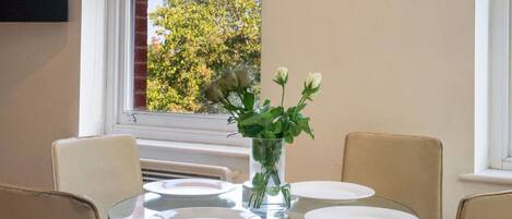 Una mesa de comedor con cubierta de vidrio y cuatro platos blancos, un florero con flores blancas, y una ventana que da a vistas de árboles.