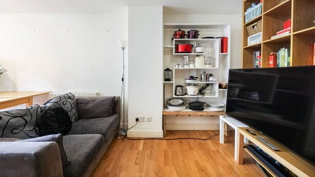 Ein Wohnzimmer mit grauem Sofa, einer TV-Konsole mit Flachbildfernseher, einem weißen Regal mit Küchenutensilien und Holzboden.