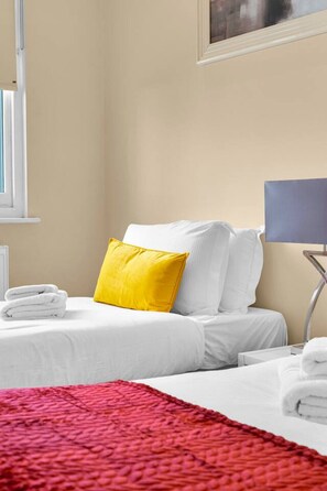A hotel room with a bed, a red blanket, a yellow pillow, white pillows, a white bedside table, and a lamp. - 2BR 2BA Kensington | Walk to High St Tube (Greater London)