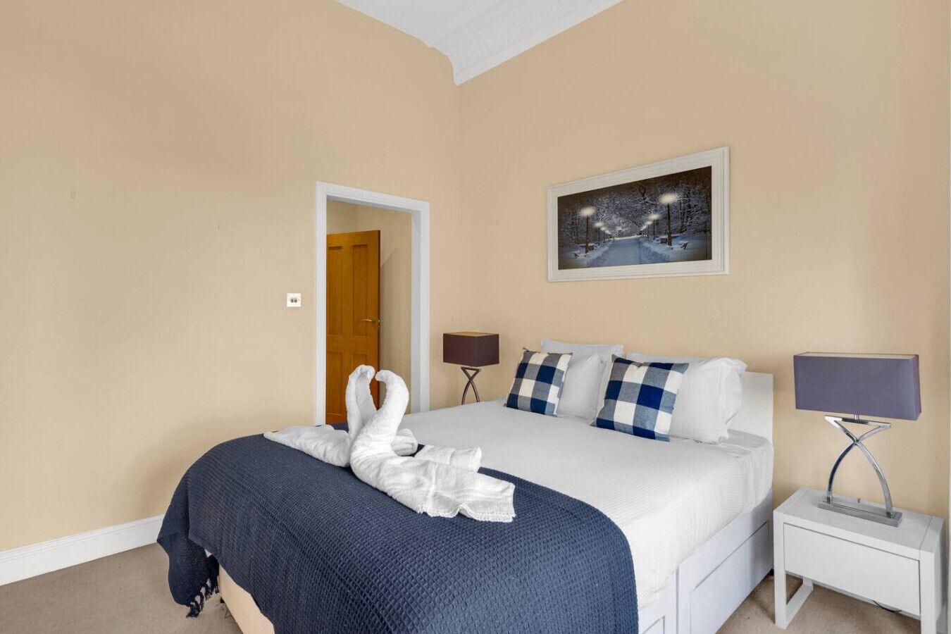 A hotel room with a bed, two bedside lamps, a framed picture on the wall, and a door leading to another room.