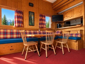 Interior de una cabaña con mesa de madera, cuatro sillas, un banco con cojines escoceses, un televisor y una zona de cocina.