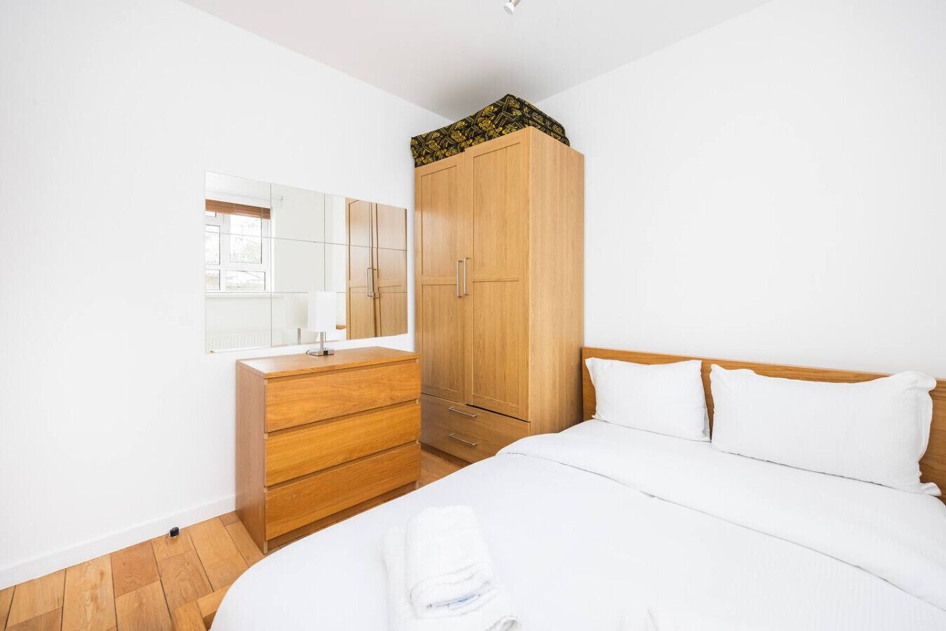 A bedroom with a bed, a wooden wardrobe, a mirror, and a window.