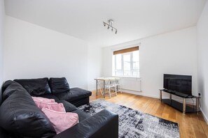 A modern living room with a black leather sofa, a small white table, a TV on a stand, and a window with blinds. - 2BR in Marylebone l Walk to Tube l Self Check-In (Greater London)