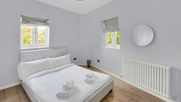 Un dormitorio con cama, dos ventanas, un radiador y un espejo redondo.
