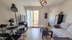 A modern living room with a sofa, a dining table with chairs, a standing fan, and a ceiling light fixture. - Studio cabin in Cap d'Agde, parking - 2 guests (Cap d'Agde)