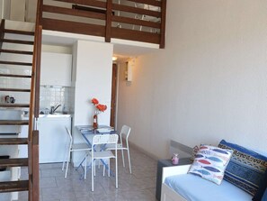 A compact living space with a kitchenette, a dining area with a table and chairs, and a bed with patterned pillows. - Studio mezzanine with sea view - 3 guests (Fleury-Saint-Pierre-la-Mer)