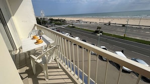 A balcony with a table and chairs, overlooking a beach and a road with parked cars. - Apartment sea view - 4 guests (Saint-Jean-de-Monts)