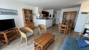 A living room with a TV, wicker chairs, a wooden coffee table, a sofa, and a kitchen area with cabinets and a dining table. - Apartment sea view - 4 guests (Saint-Jean-de-Monts)