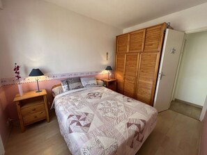 Quarto com cama, duas mesas de cabeceira, um criado-mudo, guarda-roupa e uma porta.