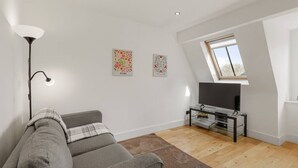 A modern living room with a grey sofa, a floor lamp, a TV on a glass stand, and two floral wall art pieces. - 1BR in Prime Islington Location near Angel Station (Greater London)