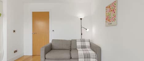 Una habitación con un sofá gris, un plaid, una lámpara de pie, una puerta de madera y una obra de arte floral en la pared.