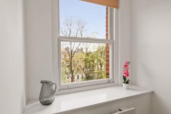 A window with a view of buildings and trees, a white radiator, and a vase with flowers on the windowsill. - 1BR in Prime Islington Location near Angel Station (Greater London)