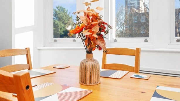 A dining table with a vase of flowers, wooden chairs, and a view of buildings through the windows. - 1BR Flat• Steps from Highbury & Islington Station (Greater London)