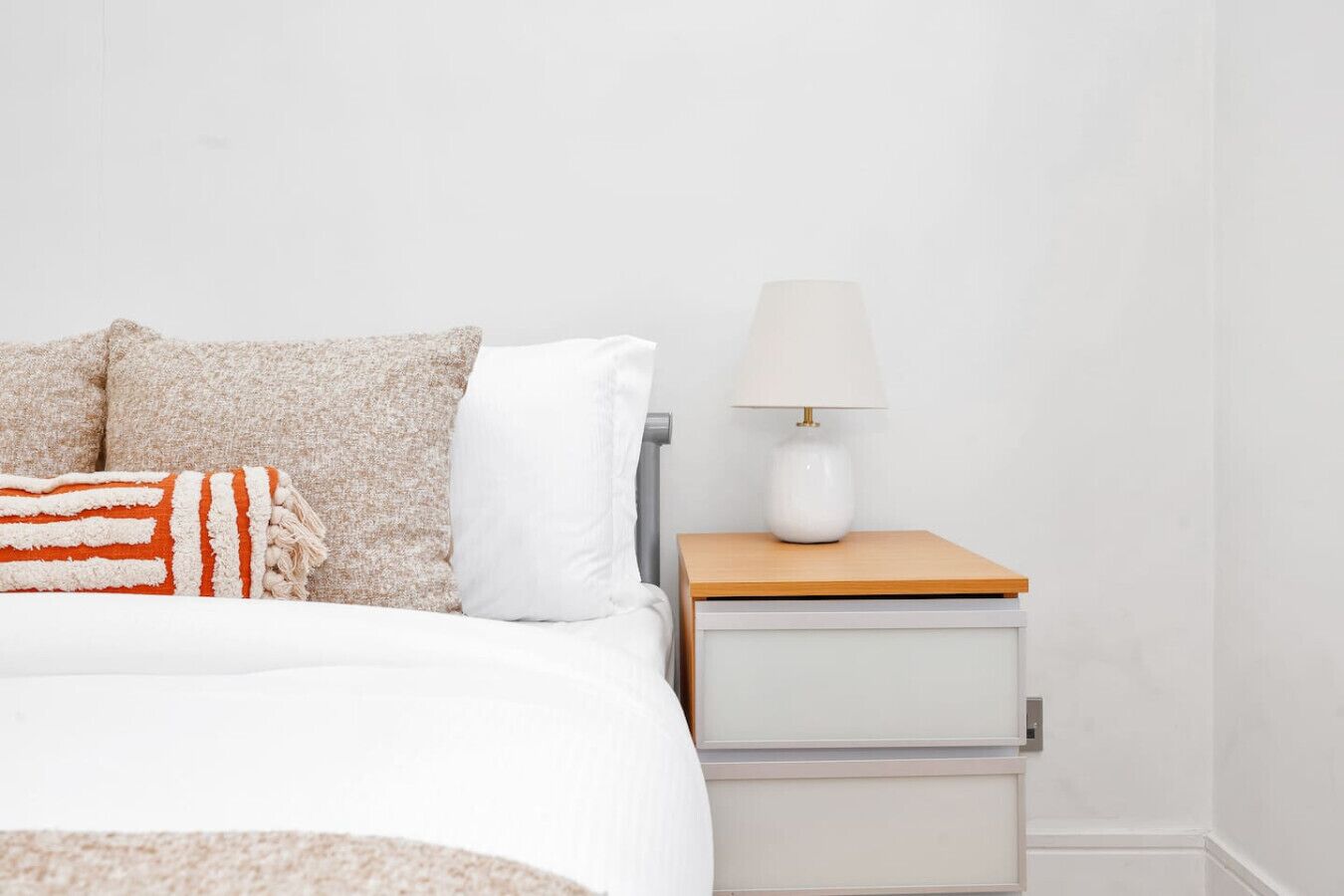 A bed with a white comforter, a beige and white pillow, and a striped blanket. There is a bedside table with a white lamp and a drawer.