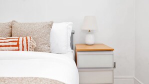 A bed with a white comforter, a beige and white pillow, and a striped blanket. There is a bedside table with a white lamp and a drawer. - 1BR Flat• Steps from Highbury & Islington Station (Greater London)