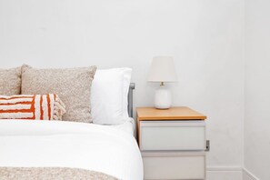 Ein Bett mit weißer Decke, einem beigen und weißen Kopfkissen und einer gestreiften Decke. Ein Nachttisch mit weißer Lampe und einer Schublade sind ebenfalls vorhanden.