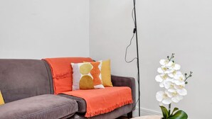 A grey sofa with a red throw blanket, a patterned cushion, and a white orchid in a vase on a round table. - 1BR Flat• Steps from Highbury & Islington Station (Greater London)