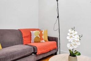 Ein graues Sofa mit roter Decke, einem gemusterten Kissen und einer weißen Orchidee in einer Vase auf einem runden Tisch.