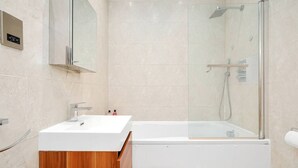 A modern bathroom with a white sink, wooden vanity, a glass-enclosed shower, and a marble-tiled wall. - 1BR Flat• Steps from Highbury & Islington Station (Greater London)