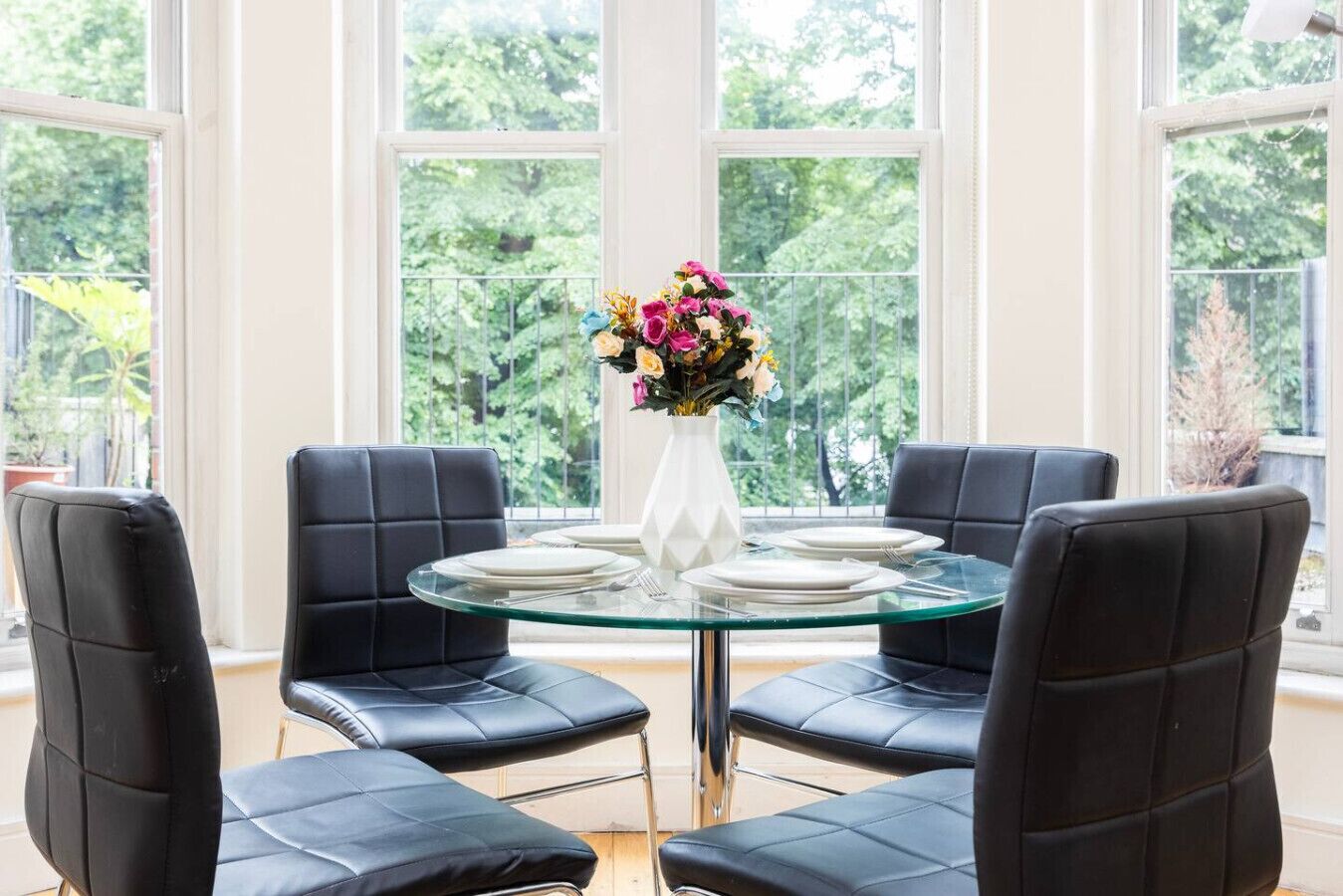A dining area with a round glass table, four black leather chairs, and a white vase with flowers.