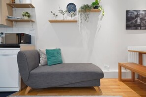 A modern living room with a grey sofa, a wooden coffee table, a kitchen area with appliances, and a wall-mounted clock. - 2-BR in the Heart of Soho - 5 guests (Greater London)