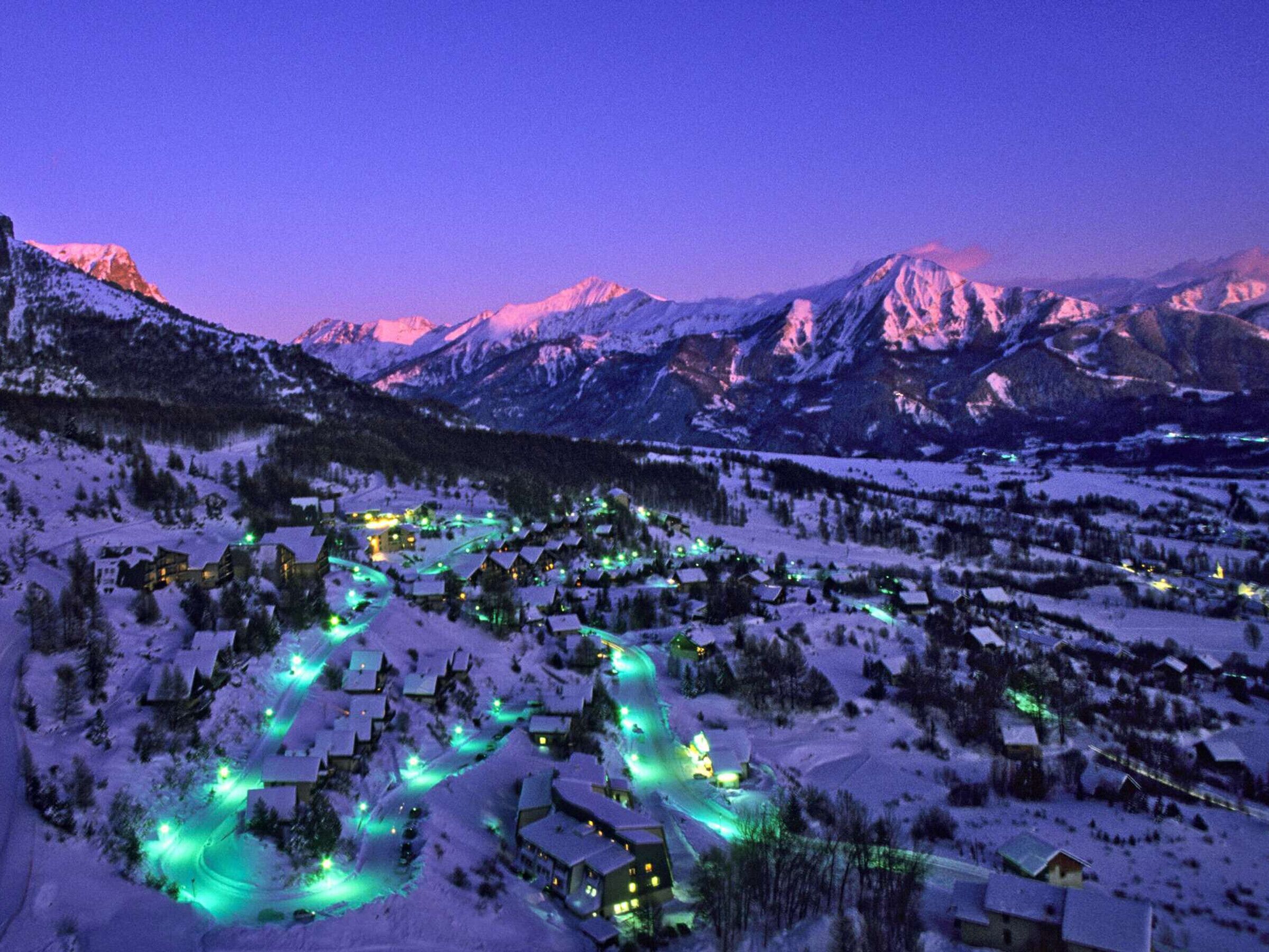 A snow-covered village with buildings illuminated by green lights, surrounded by mountains.