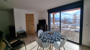 Een eetgedeelte met een tafel en stoelen, een televisie en een deur naar een balkon met uitzicht op een besneeerd landschap.