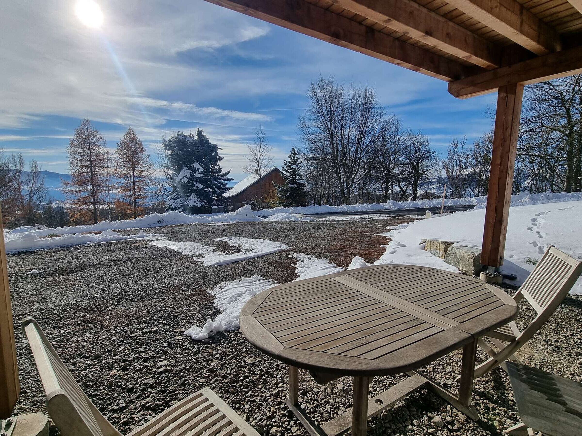 Un espace extérieur abrité, avec une table et des chaises, un chemin de gravier, un sol recouvert de neige et une vue sur des arbres et un bâtiment en arrière-plan.