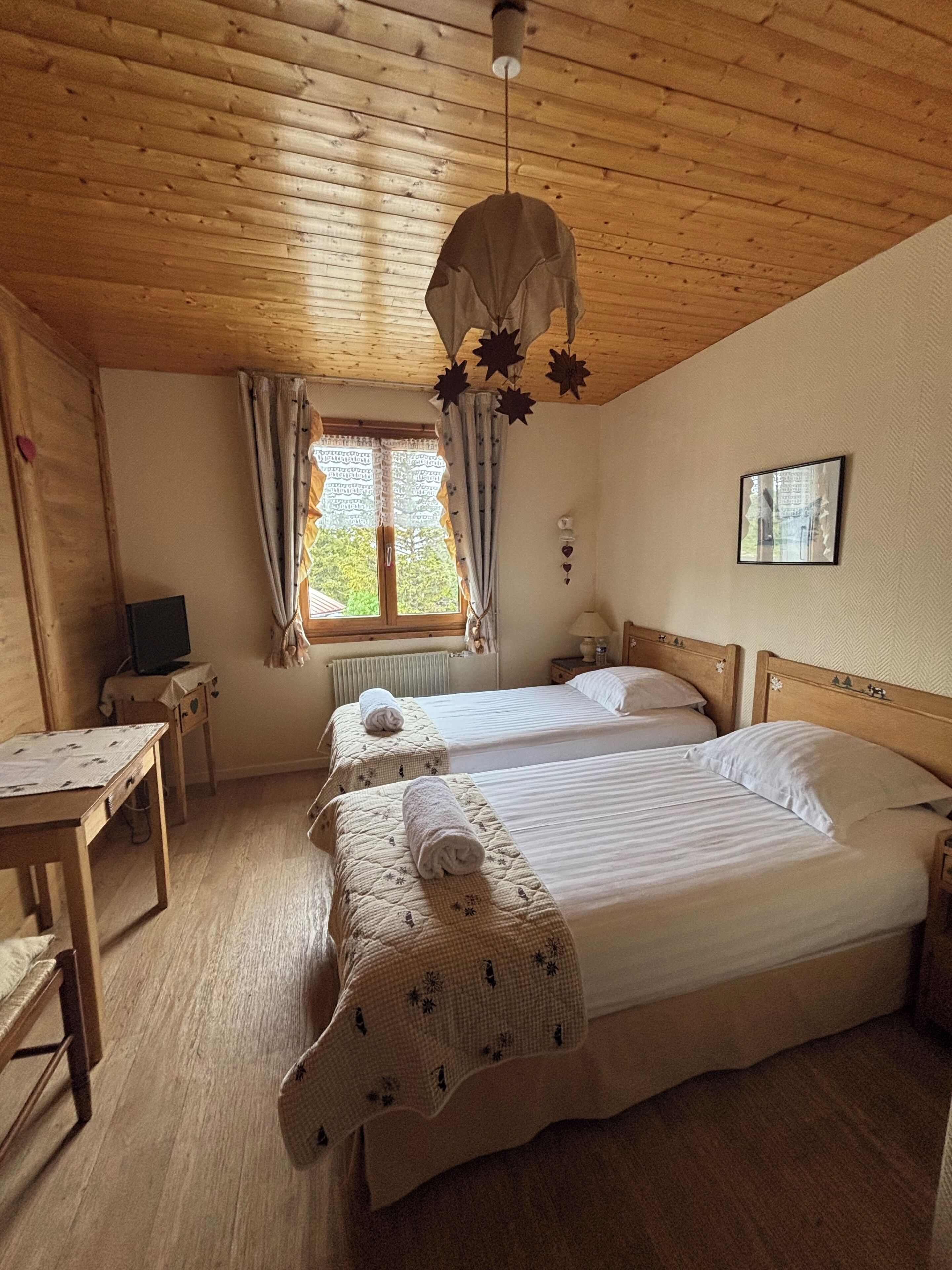 A room with two beds, a wooden ceiling, a window with curtains, a bedside table, and a small desk with a chair.