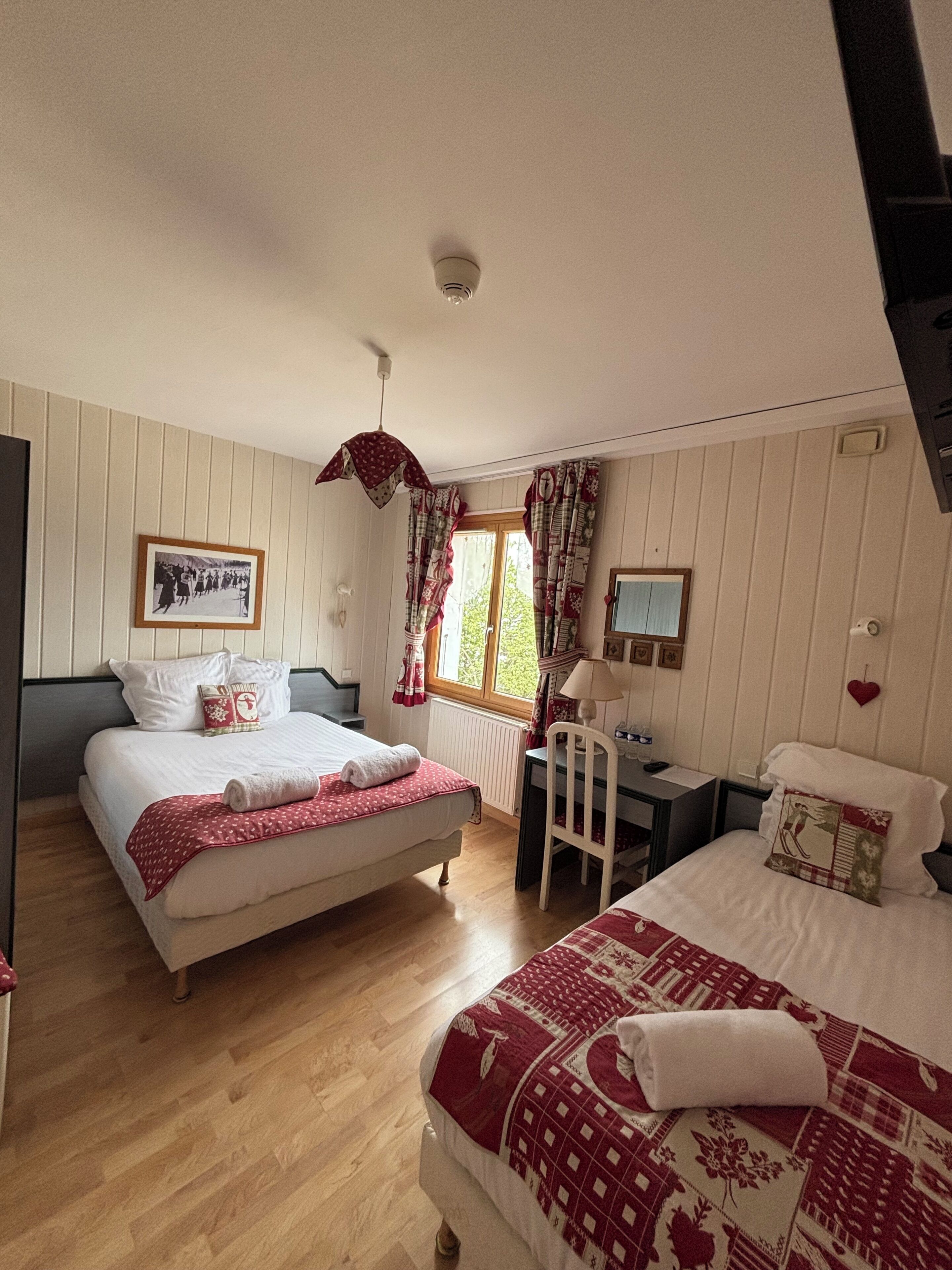 A bedroom with two beds, a desk, a chair, a window with curtains, a hanging light fixture, and a framed picture on the wall.