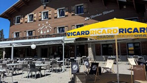 Un área de estar al aire libre con mesas y sillas, un edificio con un letrero que dice 'Les Rochers Blancs', y un toldo amarillo con la inscripción 'RICARD RICARD'.
