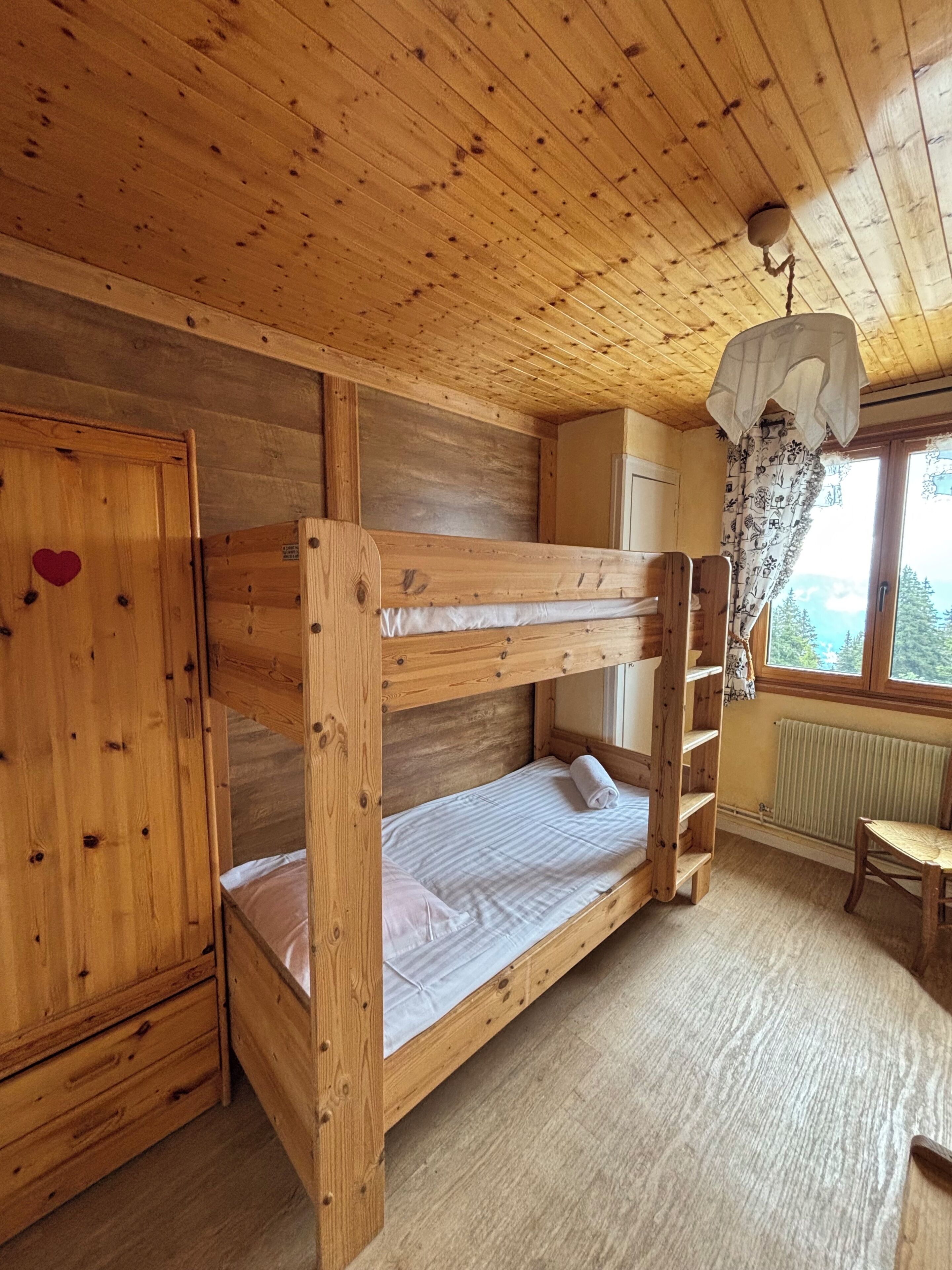 A bunk bed room with a wooden ceiling, a wardrobe with a heart decoration, a radiator, and a window with a view.