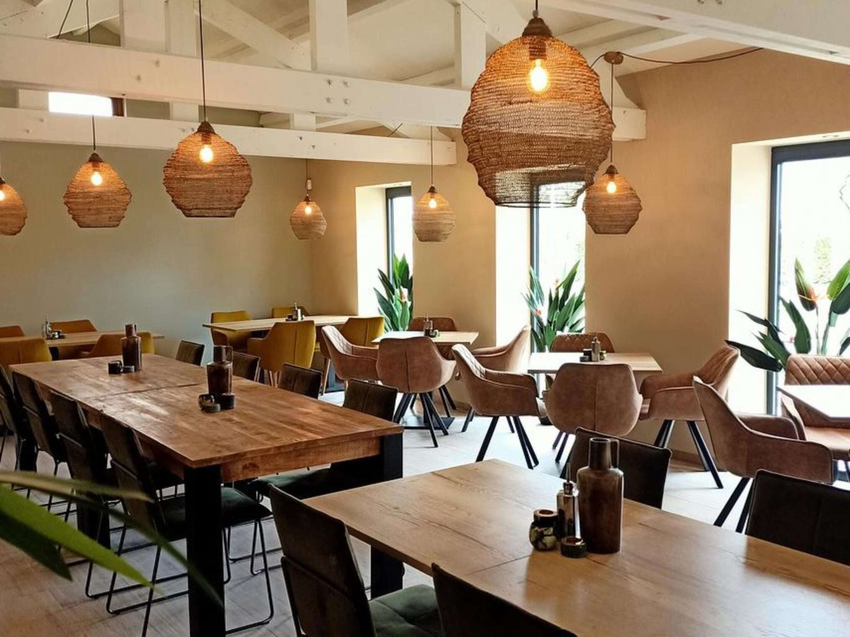 Een eetruimte met houten tafels en stoelen, geweven hanglampen en grote ramen voor natuurlijk licht.