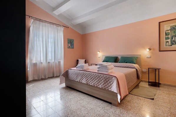 A bedroom with a bed, a nightstand, a wall-mounted lamp, a painting, and a window with curtains.
