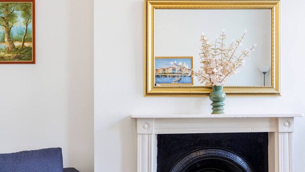 Una habitación con chimenea, un cuadro enmarcado sobre ella y un florero con flores en la repisa.