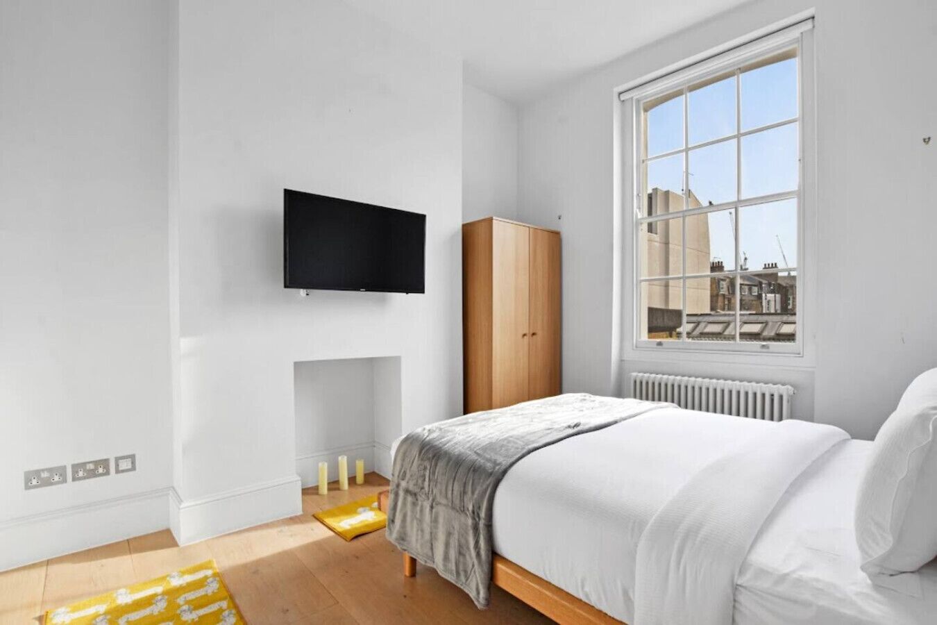 A bedroom with a bed, a TV mounted on the wall, a wardrobe, a window with a view, and a radiator.