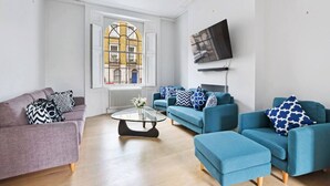 Una sala de estar moderna con un sofá gris, un juego de sofá azul, una mesa de centro de vidrio y un ventanal con vistas a edificios.