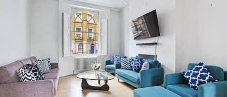 A modern living room with a grey sofa, a blue sofa set, a glass coffee table, and a large window with a view of buildings.