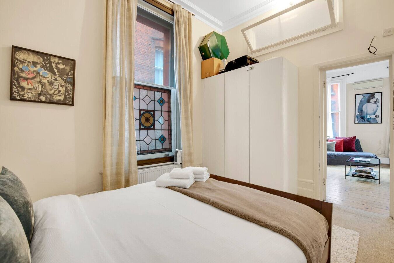 A bedroom with a bed, a window with curtains, a radiator, a wardrobe, and a painting on the wall.