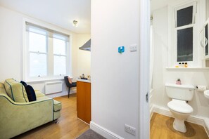 A room with a sofa, a small table, a window, a radiator, a door, and a toilet. - Stylish Studio in Leicester Square - 2 guests (Greater London)