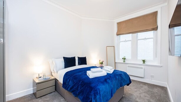 Een slaapkamer met een bed, nachtkastjes, een raam met een jaloezie en een radiator.
