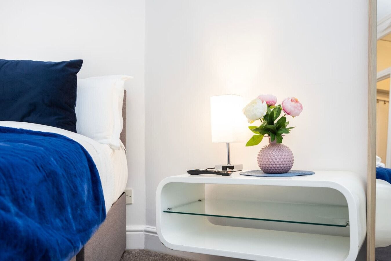 A modern hotel room with a bed, a nightstand with a lamp and vase, and a glass shelf.