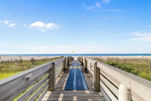 Ein hölzerner Steg führt zu einem Strand mit blauem Himmel.