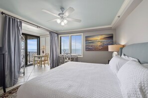 Une chambre d’hôtel avec un grand lit, un bureau avec une chaise, un balcon avec des rideaux et un ventilateur de plafond.