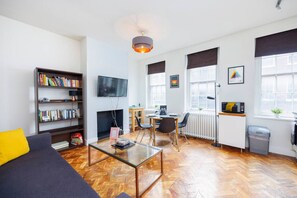 A modern living room with a sofa, a glass coffee table, a bookshelf, a fireplace, a TV, a desk with a computer, and large windows. - 1BR in Dean Street Central - 4 people (Greater London)
