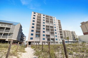 Een hotel met meerdere verdiepingen en balkons, een zandstrand op de voorgrond en een helderblauwe lucht.