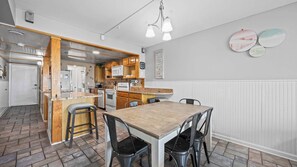 A compact kitchen with wooden cabinets, a central island, and a dining area with a table and chairs. - Castaways 1C by Liquid Life (Gulf Shores)
