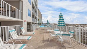 Zona junto a la piscina con sillones de descanso y sombrillas, un cartel con los horarios de cierre de la piscina y vista a la playa.