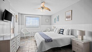 Un dormitorio con cama, ventilador de techo, televisor, ventana con persianas, una silla, mesita de noche con lámpara y cómoda.