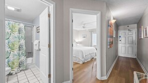 Ein Flur mit einem Badezimmer links, einem Schlafzimmer mit Bett und Deckenventilator in der Mitte und einer Tür rechts.